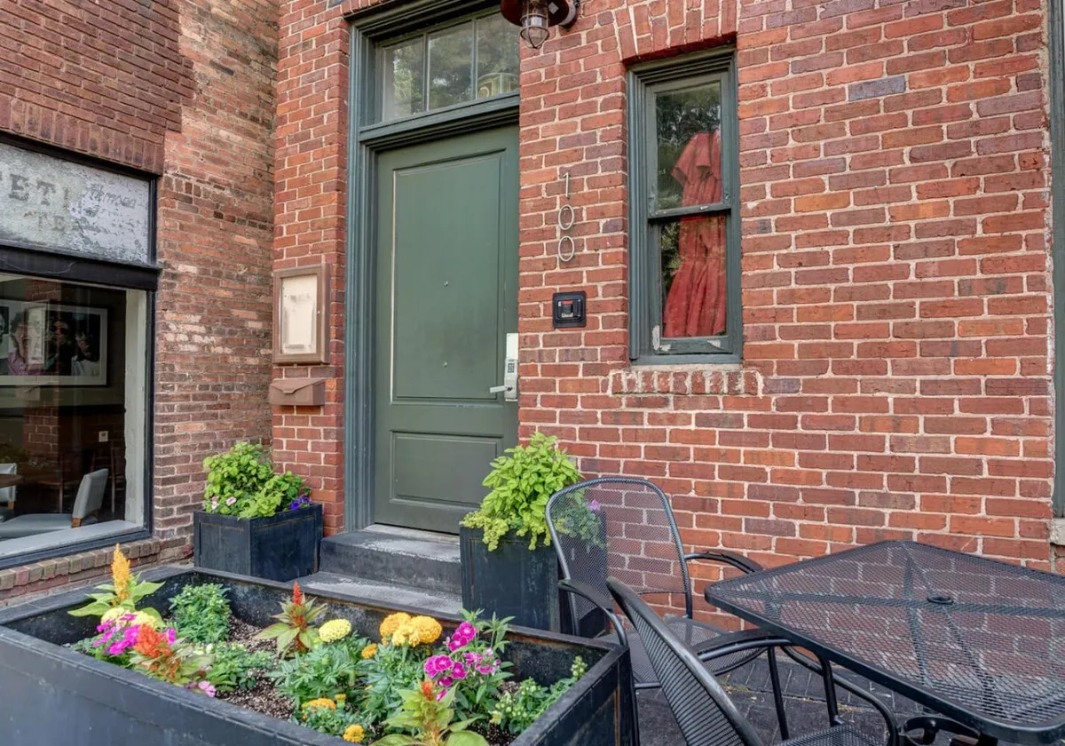 An exterior view of the building’s inviting green front door, framed by a flower bed and a window with a splash of color from a red dress. There’s a small table that hints at the possibility of relaxing outdoors or mingling with neighbors, reinforcing the building’s welcoming vibe.