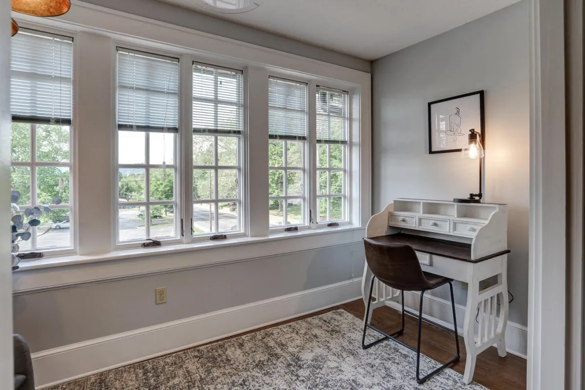 A bright and calm corner of the living area with large windows, a white desk, a decorative light fixture, and a plush rug. The windows offer urban views and fill the space with light, connecting the interior to the energy of the neighborhood.