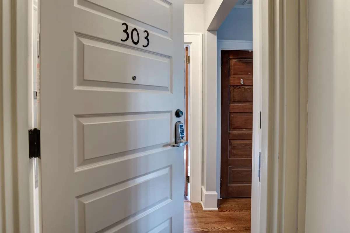 This image captures the entryway of unit 303, with the open door inviting you in and a neighboring wooden door visible to the right. The hallway suggests a communal atmosphere, where residents might cross paths and exchange greetings as they come and go.