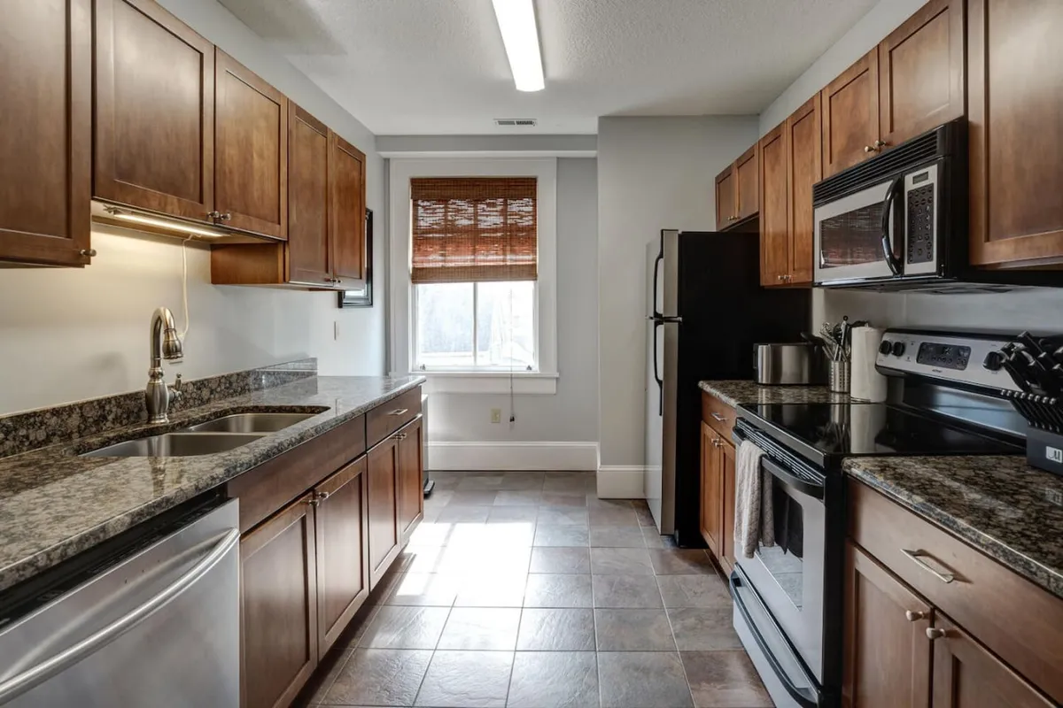 A warmly lit kitchen scene focused on granite countertops, wooden cabinets, and modern stainless steel appliances. The space feels functional and inviting, perfect for preparing meals or gathering with friends and family. Sunlight streams into the room, adding to its bright, lively ambiance.