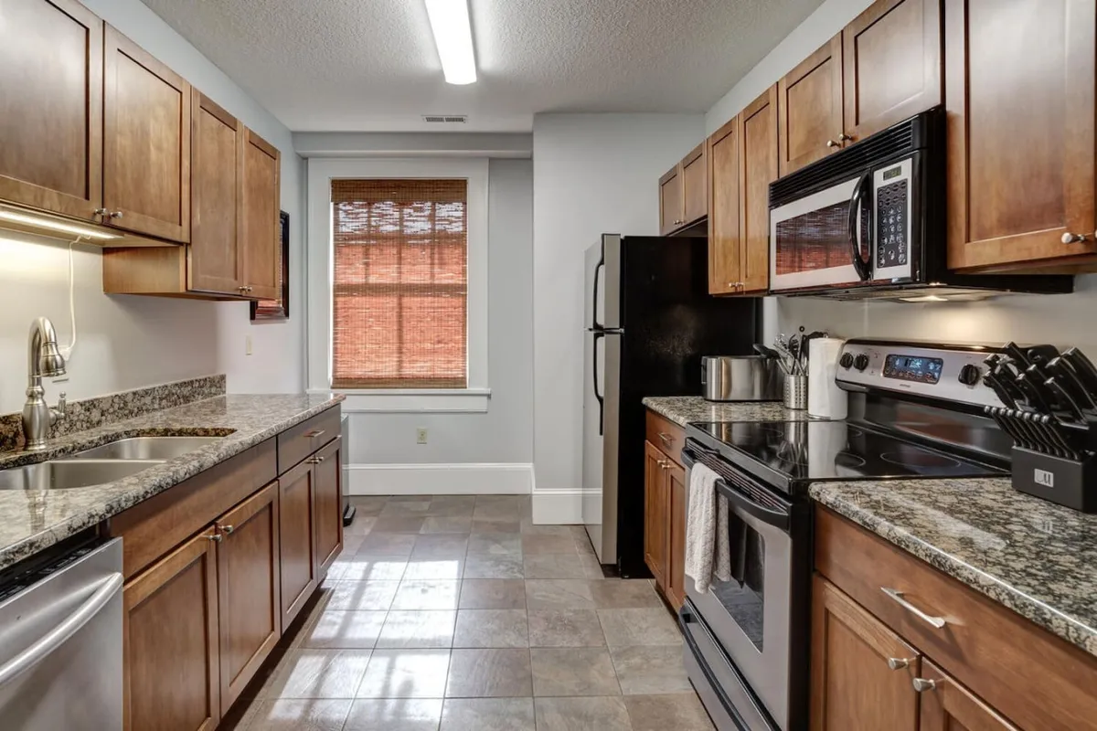 This image features a stylish, well-equipped kitchen with granite countertops, stainless steel appliances, and warm wooden cabinetry. Natural light filters in through a window with blinds, casting a welcoming glow over the space. The kitchen is clearly set up for cooking, baking, and socializing.