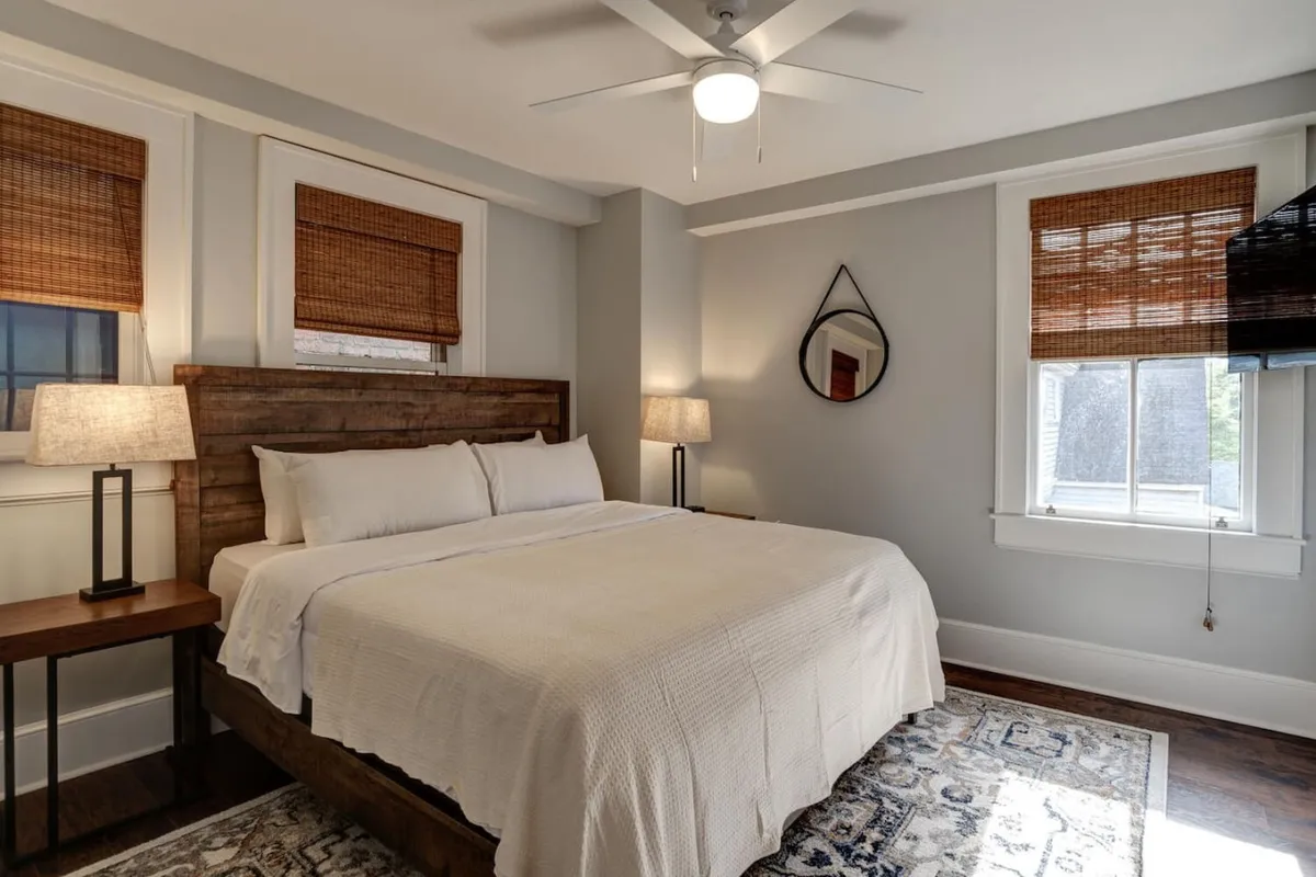 A serene bedroom featuring a comfortable bed adorned with plush bedding, flanked by matching nightstands and soft-glowing lamps. A decorative mirror and a cozy rug complete the restful ambiance, while gentle morning light streams through the unseen window.