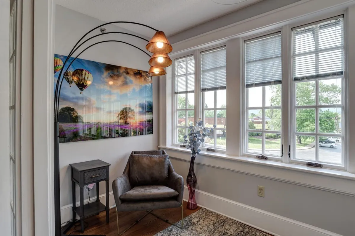 This inviting condo living space features a modern grey armchair beside an elegant arc floor lamp, complemented by vibrant local artwork and a decorative plant. Sunlight spills through large windows, creating a tranquil nook perfect for relaxing, reading, or sharing moments with friends and family.