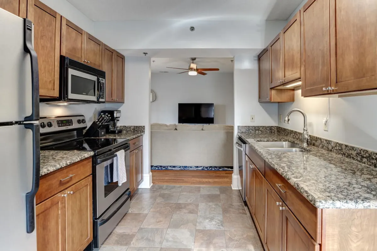 This image captures a spacious kitchen with stainless steel appliances and granite countertops. The open layout extends toward the living room, hinting at a seamless flow between cooking, dining, and social spaces—ideal for entertaining or casual gatherings.
