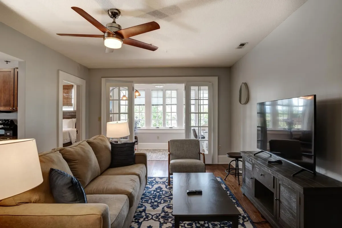 This image features a cozy living room with a comfortable sofa, a television, coffee table, a modern light fixture, and prominent windows that allow natural light to flood the space. The inviting arrangement is designed for relaxation, socializing, or unwinding after a day out.